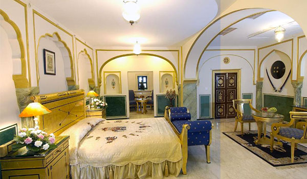 Quarto luxuoso de estilo palaciano, com cama de casal, móveis dourados, poltronas azuis, paredes brancas com detalhes dourados, arcos ornamentados e decoração tradicional indiana.