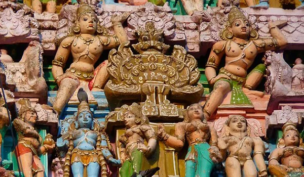 Escultura colorida e ornamentada na fachada de um templo hindu, com várias figuras divinas e humanas esculpidas. No centro há uma grande figura decorativa dourada, cercada por estátuas de diferentes personagens em poses variadas.
