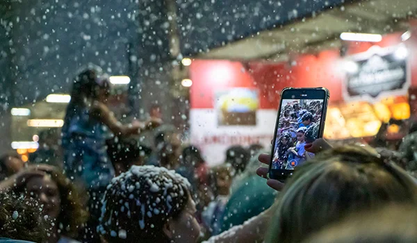 Foto de pessoas, uma com celular com neve cenográfica caindo do céu na Parada de Natal e Holambra.