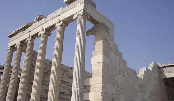 Ruínas de um antigo templo grego em Atenas, com colunas de mármore e estruturas de pedra sob um céu azul.