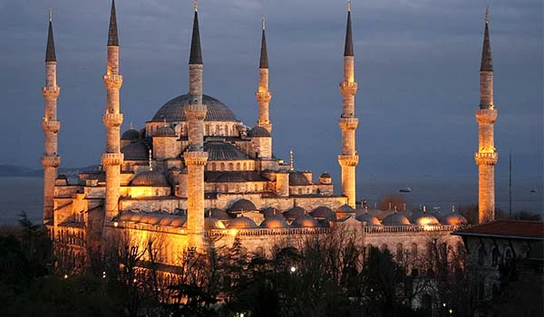 Mesquita Azul de Istambul, na Turquia, iluminada à noite, com seus minaretes imponentes e cúpulas destacadas pela iluminação dourada, sob um céu azul-acinzentado ao entardecer.
