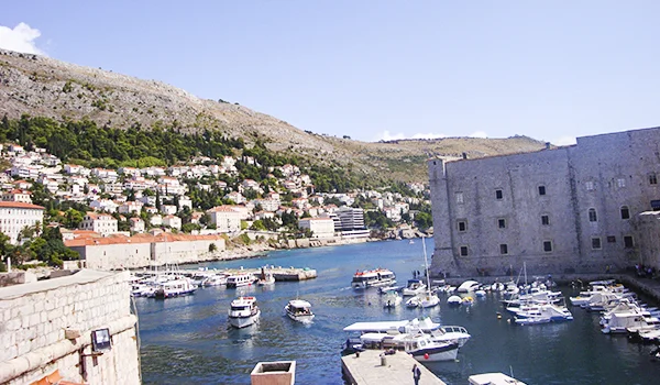 Vista de um porto costeiro com barcos atracados, muralhas históricas de pedra à direita e casas coloridas espalhadas pela encosta, cercadas por vegetação e montanhas sob um céu azul.