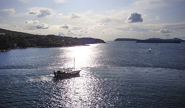 Vista panorâmica de uma baía com águas azuis e brilhantes, cercada por colinas arborizadas. Um barco à vela navega em primeiro plano, enquanto o sol reflete intensamente sobre a superfície do mar. Ao fundo, pequenas ilhas e um céu azul com nuvens dispersas compõem a paisagem.