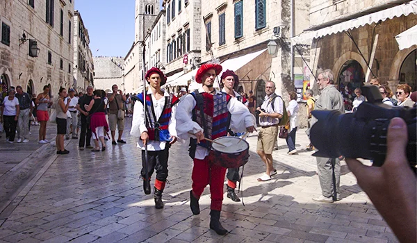 Grupo de músicos vestidos com trajes medievais, com roupas coloridas e tambores, desfila por uma rua histórica de pedra, cercada por edifícios antigos e turistas. À direita, uma pessoa fotografa a apresentação.