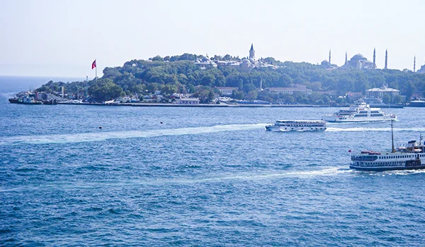 Vista do mar azul com barcos e balsas navegando pelo estreito de Bósforo, em Istambul, na Turquia. Ao fundo, uma colina arborizada abriga edifícios históricos, mesquitas com minaretes e uma bandeira turca.