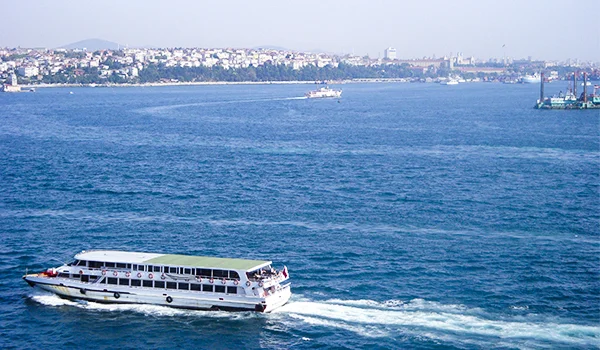 Barco de passageiros navegando em águas azuis, com a costa de uma cidade ao fundo e outras embarcações no horizonte, sob um céu claro.