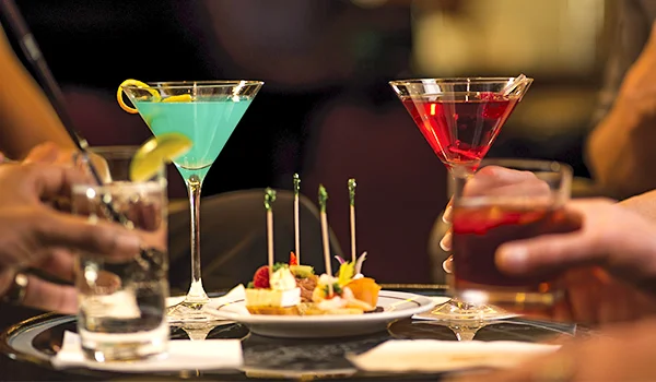 Fotografia de duas pessoas brindando com coquetéis coloridos em taças de martíni, acompanhados de aperitivos servidos em um prato sobre a mesa de um bar com iluminação aconchegante.