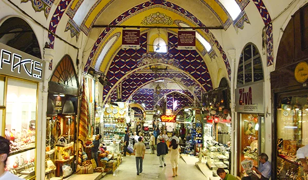 Interior do Grande Bazar de Istambul, na Turquia, com corredores repletos de lojas de joias, tapetes, luminárias e artesanatos, sob tetos abobadados decorados com mosaicos coloridos e padrões ornamentais. Turistas circulam pelo movimentado mercado tradicional.