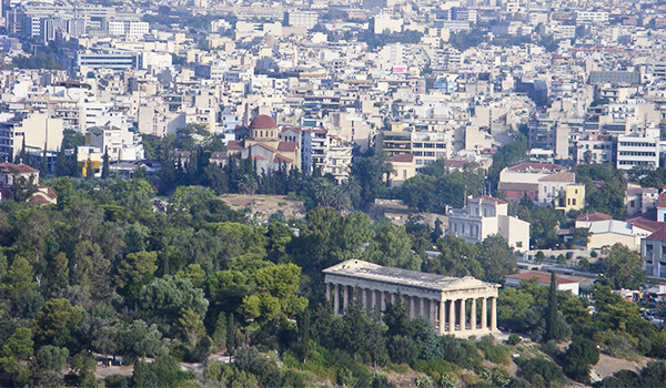 Vista panorâmica de Atenas, na Grécia, com o Templo de Hefesto em primeiro plano, cercado por árvores e vegetação, e uma extensa área urbana com edifícios brancos ao fundo.