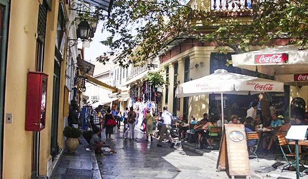 Rua estreita e movimentada em Atenas, na Grécia, com cafés e restaurantes ao ar livre, mesas e cadeiras sob guarda-sóis, lojas de artesanato e roupas nas calçadas e árvores que proporcionam sombra. Ao fundo, pedestres circulam pelo charmoso centro histórico, cercado por edifícios de arquitetura tradicional.