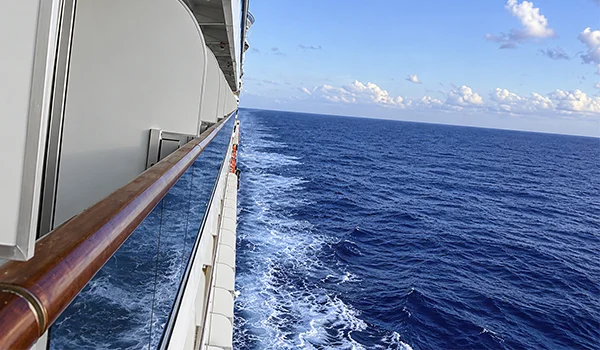 Navio de cruzeiro navegando em mar azul, visto ao longo da lateral da embarcação, com corrimão de madeira, estrutura branca, espuma das ondas e céu azul com nuvens ao fundo.