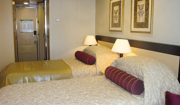 Cabine de navio de cruzeiro com duas camas de solteiro, roupas de cama estampadas, almofadas cilíndricas bordô, luminárias sobre os criados-mudos, quadros decorativos na parede e porta de entrada ao fundo.