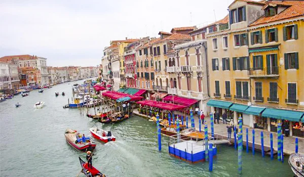 Vista do Grande Canal de Veneza, na Itália, com barcos e gôndolas navegando entre edifícios históricos coloridos, fachadas de arquitetura veneziana e restaurantes com toldos vermelhos e verdes à beira da água.