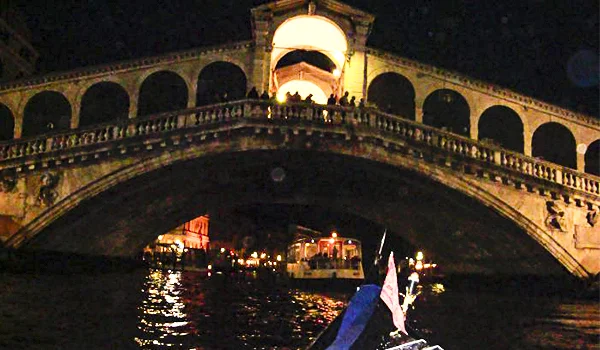 Ponte de pedra iluminada à noite sobre um canal em Veneza, com barcos navegando sob os arcos e uma gôndola em primeiro plano.
