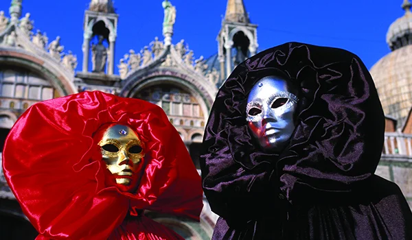 Duas pessoas usando máscaras metálicas dourada e prateada, com trajes de cetim vermelho e preto, posam em frente à Basílica de São Marcos, em Veneza, sob um céu azul.