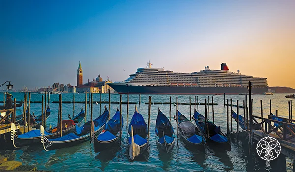 Vista de um navio de cruzeiro em Veneza ao pôr do sol, com diversas gôndolas azuis ancoradas em primeiro plano, a Basílica de São Marcos e o campanário ao fundo, sobre as águas da lagoa veneziana.