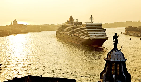 Navio de cruzeiro navegando pelo Grande Canal de Veneza, na Itália, ao pôr do sol, com a silhueta de uma estátua em primeiro plano e edifícios históricos ao fundo, iluminados pela luz dourada.