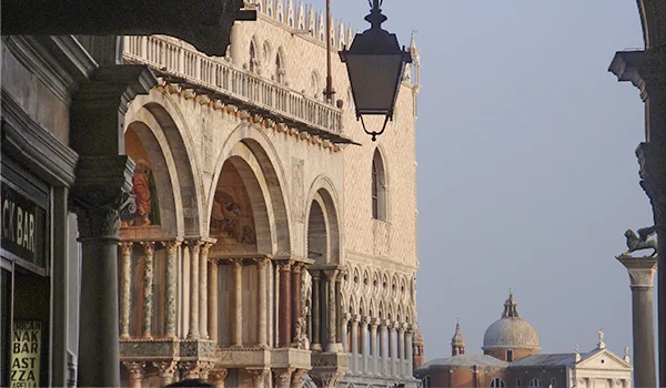 Vista de uma rua de Veneza, na Itália, com a fachada de um edifício histórico em estilo gótico, marcado por arcos, colunas e detalhes ornamentais. Ao fundo, destacam-se a cúpula de uma igreja e o céu azul, enquanto um lampião suspenso e as construções laterais emolduram a cena.
