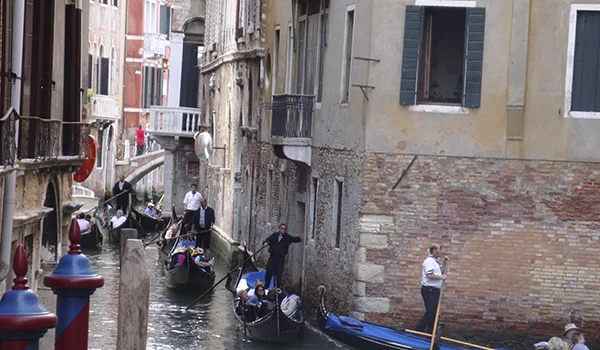 Canal estreito em Veneza, cercado por edifícios antigos, com pessoas navegando em gôndolas e barcos pela água.