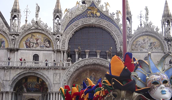Fachada ornamentada da Basílica de São Marcos, em Veneza, na Itália, com esculturas, mosaicos e arcos góticos. Na parte inferior da imagem, pessoas fantasiadas com máscaras coloridas e elaboradas celebram o tradicional Carnaval de Veneza.
