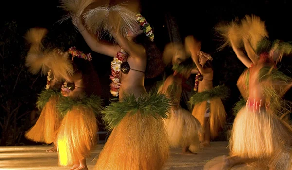 Dançarinas realizam uma apresentação de dança tradicional polinésia, usando saias de fibras naturais, adornos florais e folhas verdes. Elas movimentam os braços e os quadris em um palco iluminado, em uma apresentação noturna.