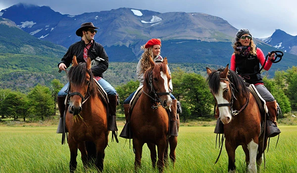 Três pessoas passeando a cavalo em uma paisagem natural com montanhas ao fundo, em um dia ensolarado, desfrutando de um passeio ao ar livre.