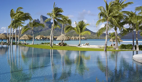 Imagem de um resort tropical à beira-mar, com uma grande piscina de borda infinita em primeiro plano, coqueiros, praia de areia branca e bangalôs sobre a água. Ao fundo, águas azul-turquesa e uma montanha verde e imponente sob um céu azul com algumas nuvens.