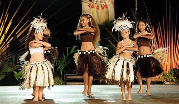 Quatro dançarinas se apresentam em um palco, usando trajes tradicionais com saias de fibras naturais, adornos de penas e cocares. Elas estão em diferentes posições de dança, diante de um cenário decorado com plantas e elementos tropicais.