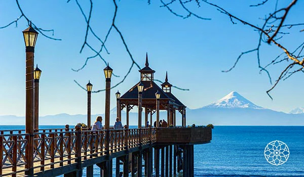 Cenário com vista para o Lago Llanquihue e o vulcão Osorno, mostrando um píer de madeira com lanternas e turistas apreciando a paisagem na cidade de Puerto Varas, na região dos Lagos, Chile.