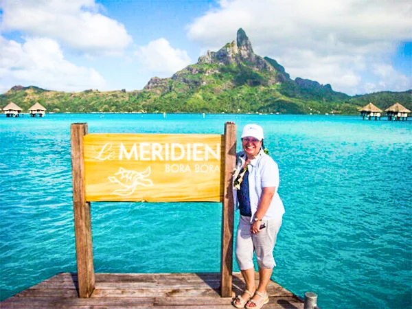 Deusa Rodrigues em um píer sobre águas azul-turquesa, ao lado de uma placa de madeira com a inscrição “Le Méridien Bora Bora”. Ao fundo, há bangalôs sobre a água e uma montanha verde e imponente sob um céu parcialmente nublado. 