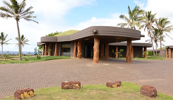 Imagem de uma construção moderna com estrutura de madeira e cobertura arredondada, rodeada por árvores de palmeira e um espaço amplo de estacionamento ao ar livre, em um ambiente tropical ao ar livre.