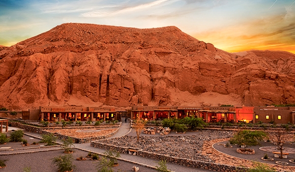 Imagem de um hotel no Deserto do Atacama ao entardecer, destacando as complexas formações rochosas e o céu colorido ao fundo, com iluminação suave nas áreas inferiores.