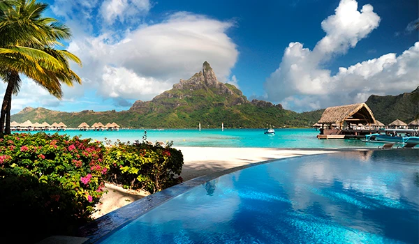 Praia paradisíaca em Bora Bora, com água azul-turquesa cristalina, piscina de um resort em primeiro plano, palmeiras e flores tropicais à esquerda, bangalôs sobre a água à direita e o imponente Monte Otemanu ao fundo, sob um céu azul com nuvens brancas.