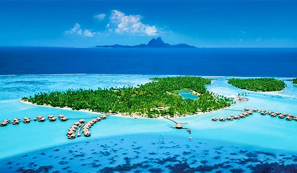 Vista aérea de uma ilha tropical cercada por águas cristalinas azul-turquesa, com vegetação verde exuberante, praias de areia clara e bangalôs sobre a água. Ao fundo, vê-se o oceano e uma cadeia de montanhas sob um céu azul com nuvens.