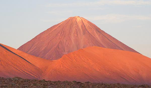 Imagem de um vulcão com pico em erupção, com encostas de cinza e lava, sob um céu parcialmente nublado. Perfeito para quem busca fotos de vulcões ativos para conteúdo de viagens ou geologia. Chile pontos turísticos.