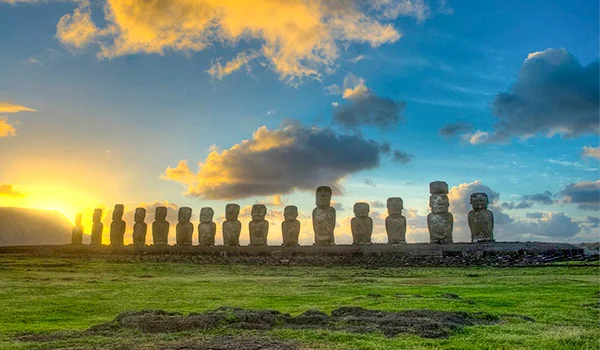 Estátuas moai de pedra alinhadas em uma paisagem verde da Ilha de Páscoa, diante de um céu azul com nuvens iluminadas pelo pôr do sol.