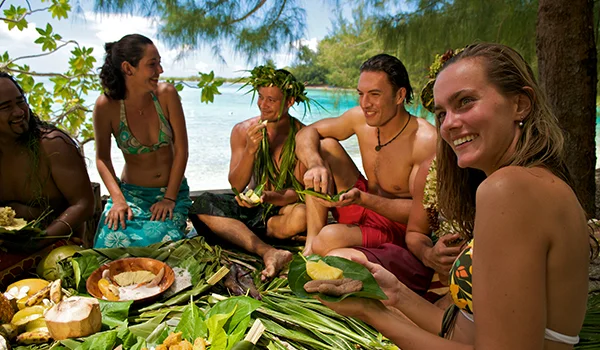 Grupo de pessoas reunido ao ar livre, à sombra de árvores, junto a águas cristalinas. Elas estão sorrindo e preparando ou compartilhando alimentos tradicionais servidos sobre folhas grandes, em um ambiente tropical.
