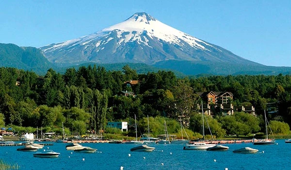 Vista panorâmica Lago Vilarrica ao fundo, com águas do lago e barcos ao primeiro plano, cercado por uma natureza exuberante e cidade à beira do lago. Lua de Mel no Chile.