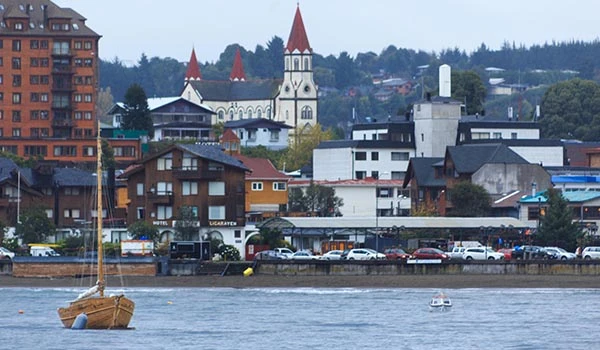 Vista do centro de uma cidade com igreja de torres vermelhas, edifícios residenciais e comerciais ao lado de um rio, com um barco de madeira ao fundo.