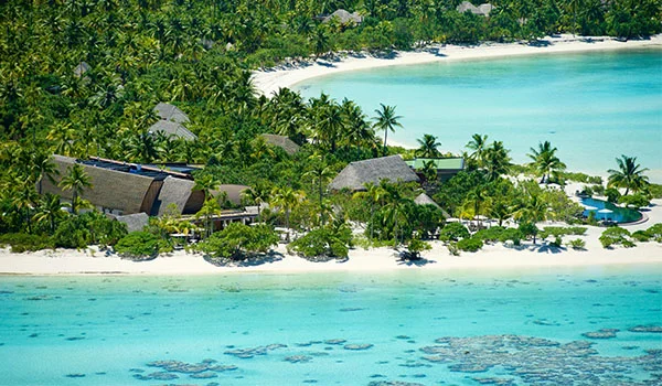 Ilha tropical com vegetação exuberante, palmeiras, bangalôs à beira-mar e uma praia de areia branca cercada por águas cristalinas em tons de azul-turquesa.