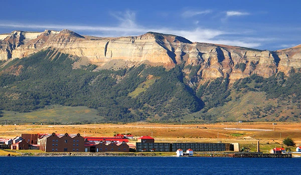 Paisagem com montanhas ao fundo, uma área industrial e um rio em primeiro plano, sob céu claro com algumas nuvens.