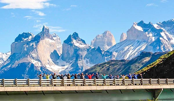 Grupo de pessoas participando de uma caminhada em uma ponte na frente de uma paisagem de montanhas nevadas e picos rochosos durante o dia. Chile pontos turísticos.