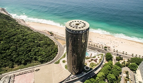 Vista aérea de um prédio alto na Praia de Copacabana, Rio de Janeiro, com mar azul e faixa de areia, cercado por vegetação e cidade.