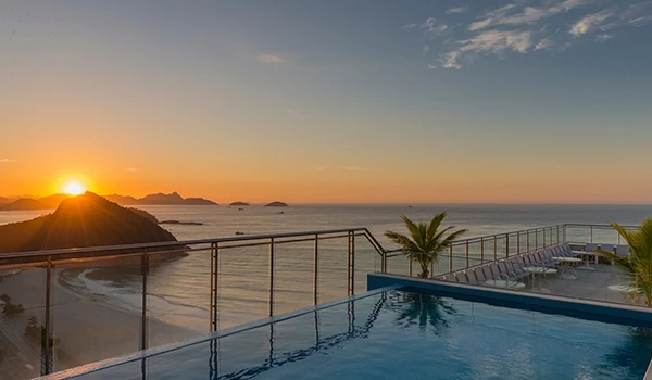 Vista de um pôr do sol na praia com piscina ao fundo, com vista para o mar e ilhas ao longe, criando uma cena tranquila e paradisíaca.