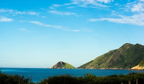 Paisagem de praia com mar azul, colinas verdes ao fundo e céu ensolarado, ideal para apreciadores de natureza e tranquilidade no litoral.