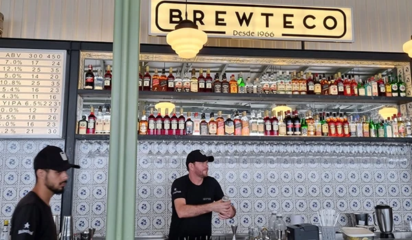 Interior de uma loja de bebidas chamada Brewteco, com prateleiras cheias de garrafas de bebidas alcoólicas, duas pessoas atendendo no balcão, decoração vintage e iluminação acolhedora.