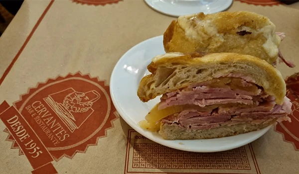 Sanduíche de pernil com pão integral e ingredientes chegado à mesa em um restaurante, com impressão de logotipo e gráficos de apostas ao fundo.