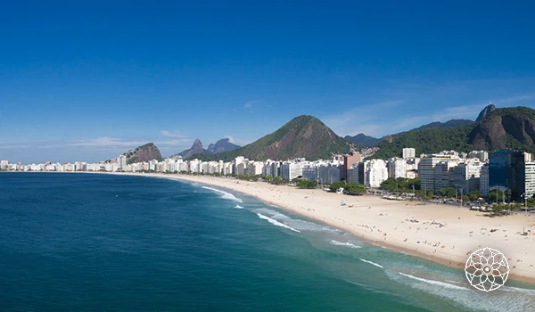 Vista panorâmica da orla da praia de Copacabana no Rio de Janeiro, com areias douradas, mar azul e as famosas montanhas ao fundo, cenário icônico do Brasil