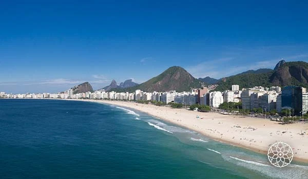 Vista panorâmica da Praia de Copacabana no Rio de Janeiro, com areia branca, mar azul e as montanhas ao fundo, em um dia ensolarado.