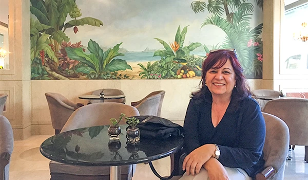 Deusa Rodrigues sentada em mesa de restaurante com ambiente tropical ao fundo, decorada com plantas e uma pintura de cenário natural na parede. Rio de Janeiro os melhores passeios.